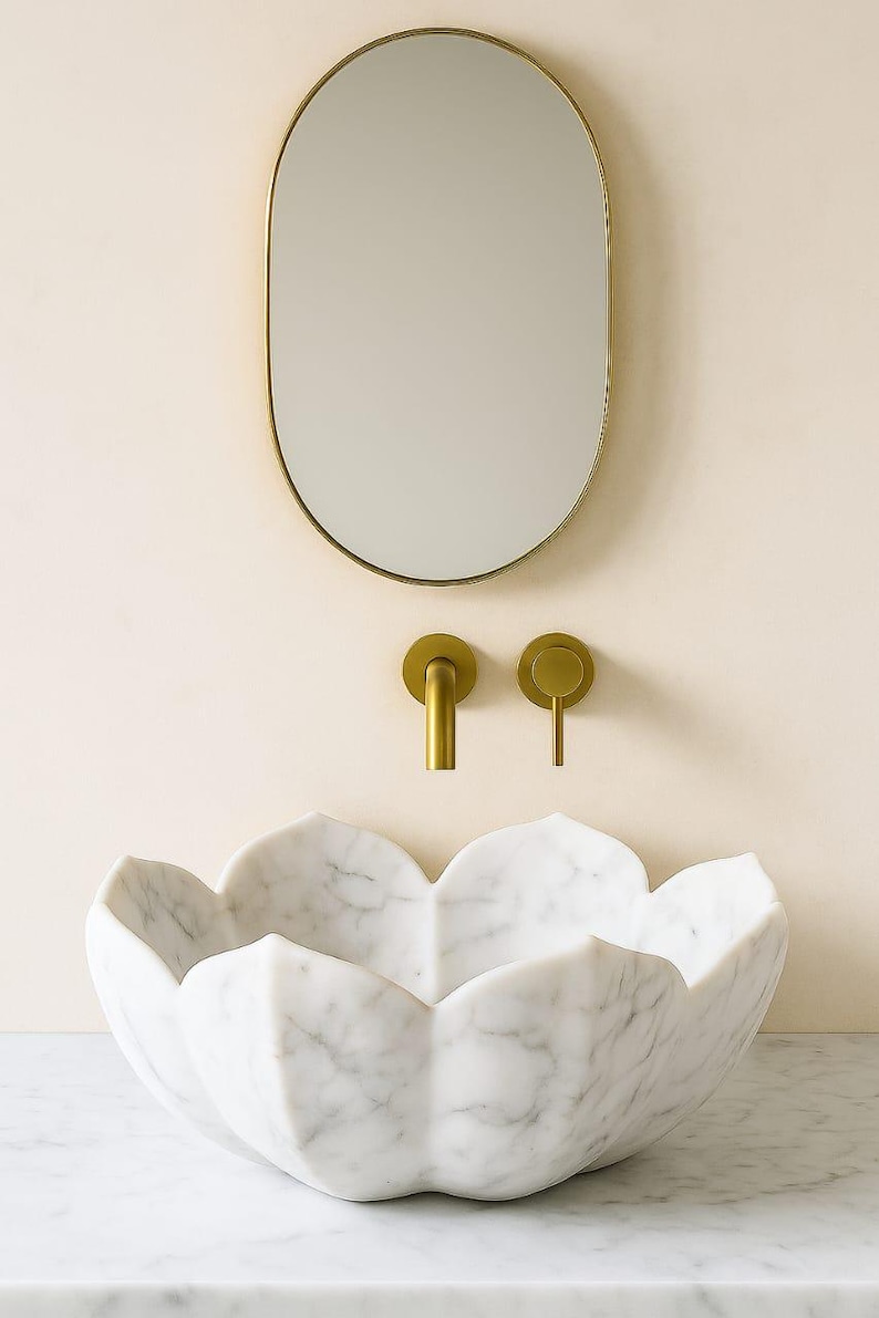May include: A white marble lotus-shaped sink with a gold tap and matching fittings. An oval gold-framed mirror hangs above the sink. The sink is set against a light beige wall.