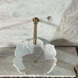 May include: A white marble flower-shaped sink with a gold tap. The sink has a central drain and is set against a textured, light-coloured wall. The tap is a simple, elegant design.