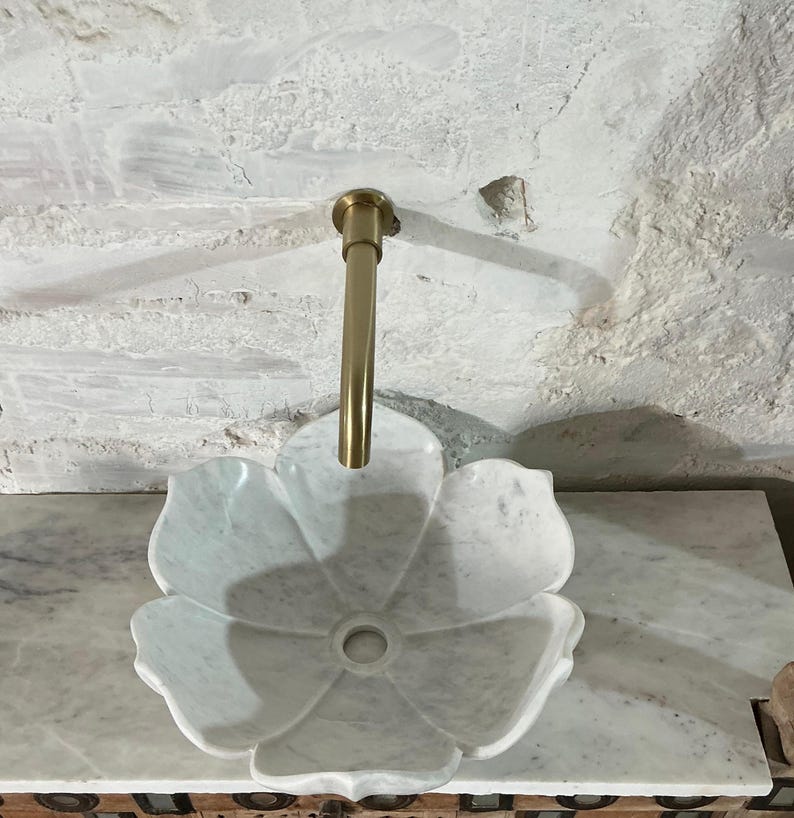 May include: A white marble flower-shaped sink with a gold tap. The sink has a central drain and is set against a textured, light-coloured wall. The tap is a simple, elegant design.