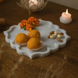 May include: A white marble serving tray with a scalloped edge holds orange sweets, marigold flowers, and almonds. A lit candle sits on the tray, and a lotus-shaped candle holder with a lit candle is in the background. The tray is on a wooden surface.