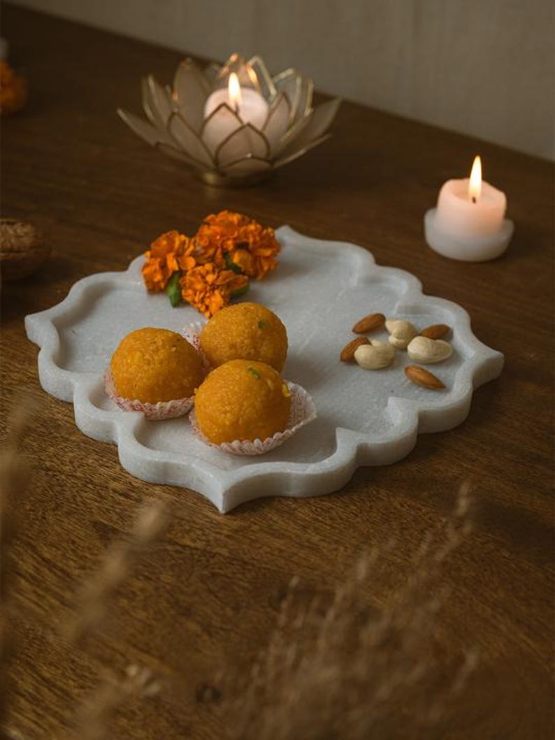 May include: A white marble serving tray with a scalloped edge holds orange sweets, marigold flowers, and almonds. A lit candle sits on the tray, and a lotus-shaped candle holder with a lit candle is in the background. The tray is on a wooden surface.