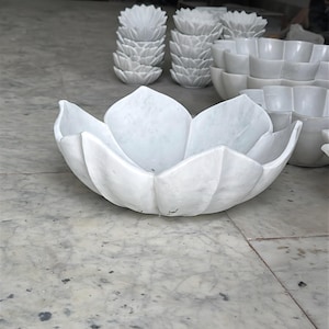 May include: A selection of white marble bowls and decorative items. The image features a large, open bowl in the shape of a lotus flower. Other pieces include smaller lotus-shaped bowls and decorative items, all in a clean, minimalist style.
