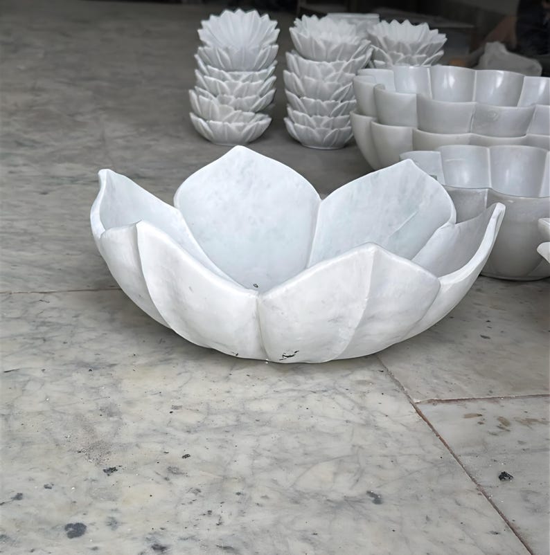 May include: A selection of white marble bowls and decorative items. The image features a large, open bowl in the shape of a lotus flower. Other pieces include smaller lotus-shaped bowls and decorative items, all in a clean, minimalist style.