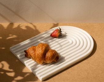 May include: A white, arched serving tray with a ridged design, holding a golden-brown croissant and a fresh red strawberry. The tray sits on a tan surface, with shadows from a plant in the background.