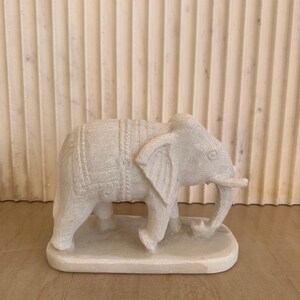 May include: A carved, off-white elephant figurine. The elephant has detailed carvings on its body and is standing on a rectangular base. The figurine is made of a stone-like material and is displayed against a neutral background.