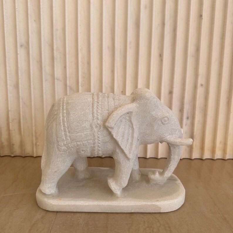 May include: A carved, off-white elephant figurine. The elephant has detailed carvings on its body and is standing on a rectangular base. The figurine is made of a stone-like material and is displayed against a neutral background.