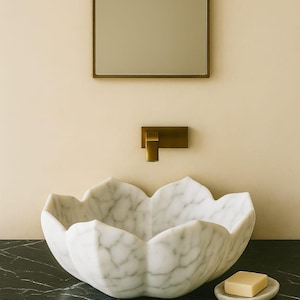 May include: A white marble lotus-shaped sink with grey veining sits on a black countertop. A small soap dish with a bar of soap is to the right. A gold-toned tap and mirror are mounted on the wall above.