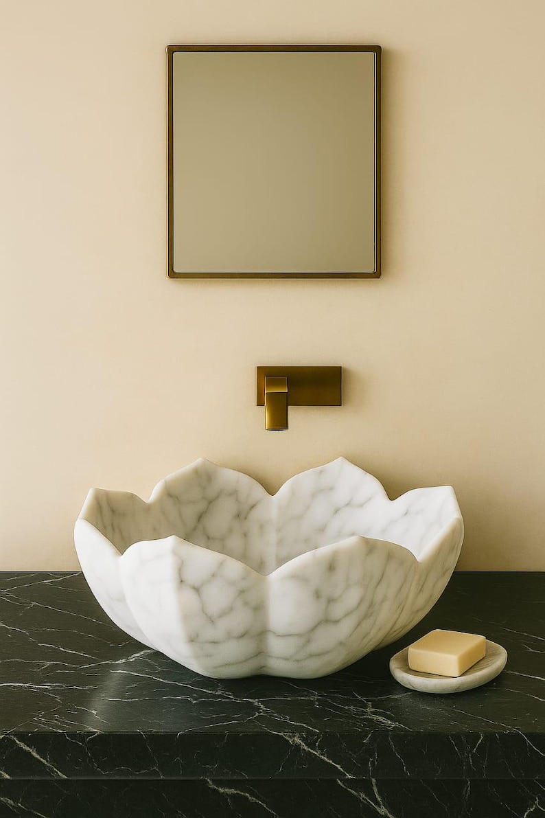 May include: A white marble lotus-shaped sink with grey veining sits on a black countertop. A small soap dish with a bar of soap is to the right. A gold-toned tap and mirror are mounted on the wall above.