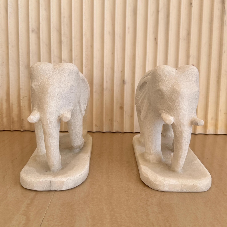 May include: Two off-white elephant sculptures. Each elephant has a detailed trunk, ears, and tusks. The elephants stand on a rectangular base. The sculptures are made of a stone-like material.