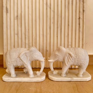 May include: Two off-white marble elephant sculptures. Each elephant has detailed carvings, including a saddle and trunk. The sculptures are on rectangular bases, and the background features a textured, vertical-lined wall.