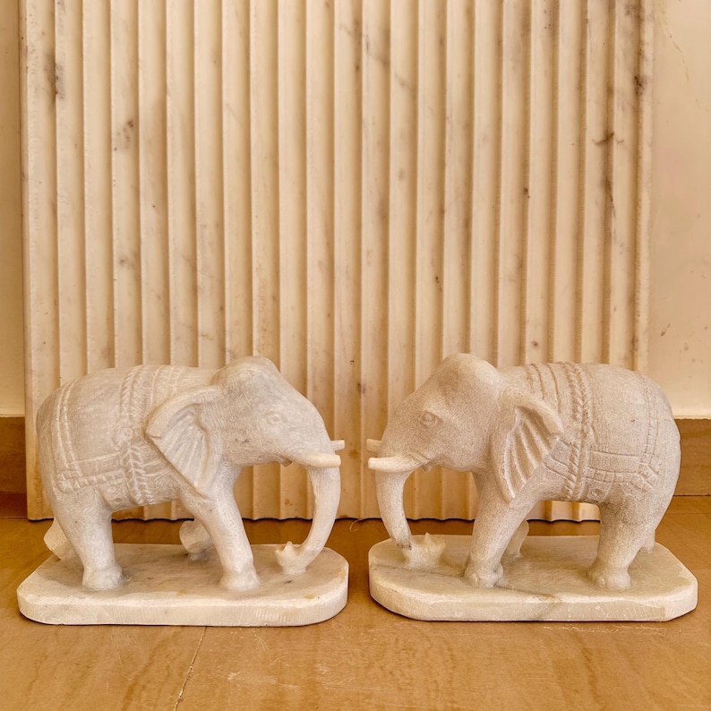 May include: Two off-white marble elephant sculptures. Each elephant has detailed carvings, including a saddle and trunk. The sculptures are on rectangular bases, and the background features a textured, vertical-lined wall.