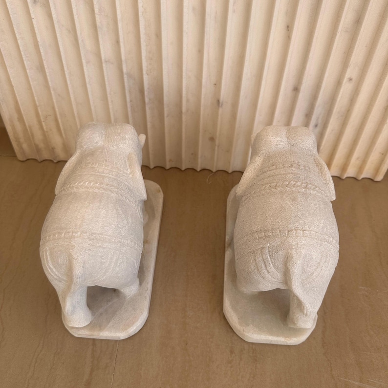 May include: Two white marble elephant sculptures, each with detailed carvings, stand on rectangular bases. The elephants face away from the viewer, showcasing their textured backs and intricate designs. The sculptures are placed on a light brown surface.