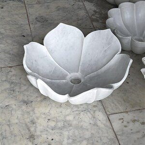 May include: A white marble sink sculpted to resemble a flower. The sink features a central drain and petal-like edges. The sink is made of white marble and is placed on a marble surface.