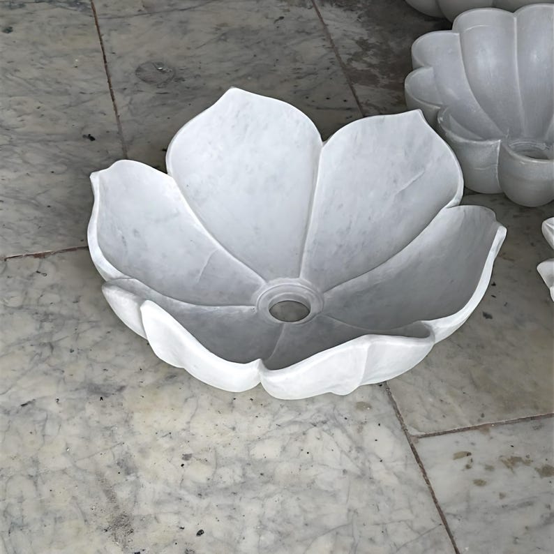 May include: A white marble sink sculpted to resemble a flower. The sink features a central drain and petal-like edges. The sink is made of white marble and is placed on a marble surface.