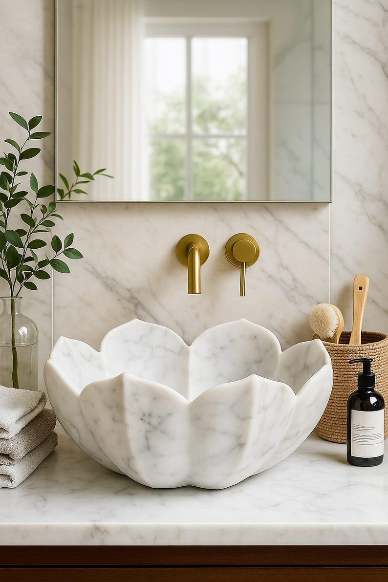 May include: A white marble lotus-shaped sink with gold taps. The sink is the focal point, set against a marble wall and countertop. A mirror, greenery, and a woven basket with toiletries complete the bathroom scene.