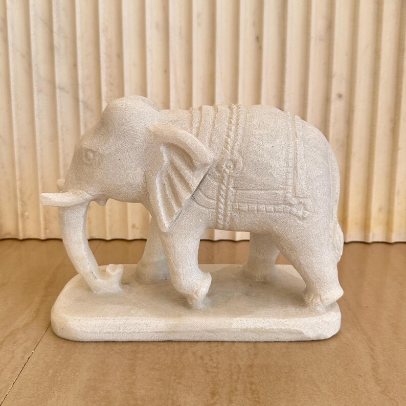 May include: A detailed, off-white elephant figurine. The sculpture features intricate carvings, including a saddle-like design on its back. The elephant stands on a rectangular base, showcasing a classic, decorative style. The figurine is made of a stone-like material.