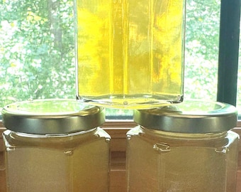 May include: Three clear glass hexagonal jars filled with a translucent golden liquid. Each jar features a metallic silver screw-top lid. The jars are arranged in a pyramid formation, with one jar positioned centrally above two others on a flat surface.