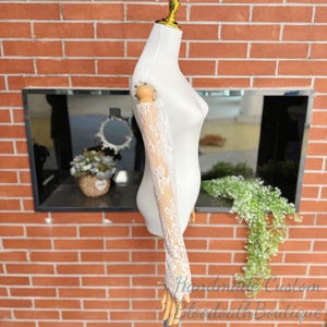 May include: A white lace sleeve displayed on a mannequin. The lace has a floral pattern and extends from the upper arm to the hand. The mannequin is set against a brick wall background.