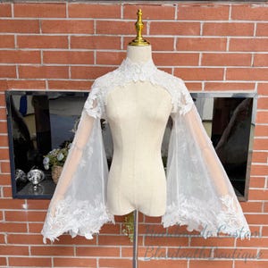 May include: A white lace bridal cape with long, flared sleeves. The cape features a high, floral-patterned collar and delicate lace detailing along the edges. The garment is displayed on a mannequin against a brick wall background.