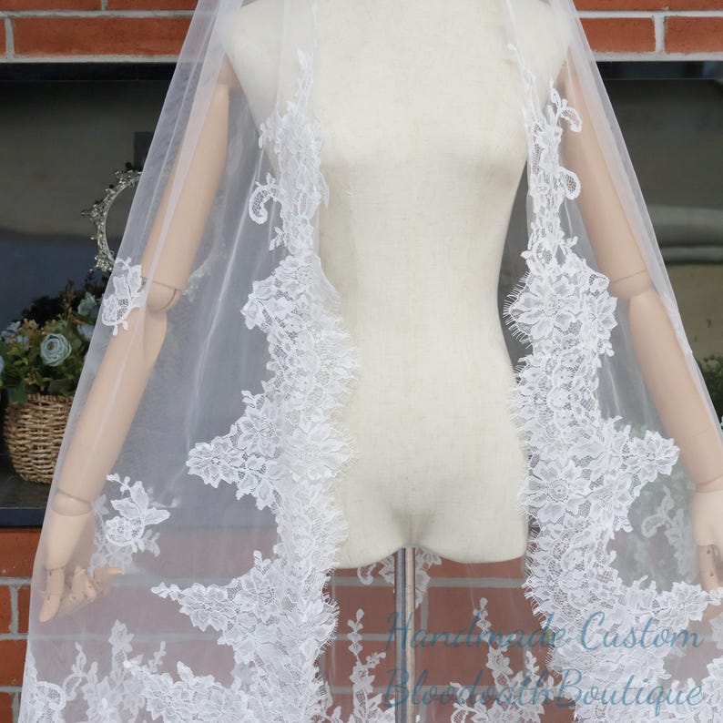 May include: A white bridal veil with intricate floral lace detailing along the edges. The veil is sheer, allowing light to pass through, and is displayed on a mannequin. The lace features delicate patterns and adds a touch of elegance.
