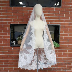 May include: A white bridal veil with intricate lace detailing along the edges. The veil is displayed on a mannequin against a brick wall background. The lace design adds a delicate and elegant touch to the wedding accessory.