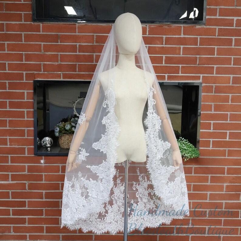 May include: A white bridal veil with intricate lace detailing along the edges. The veil is displayed on a mannequin against a brick wall background. The lace design adds a delicate and elegant touch to the wedding accessory.