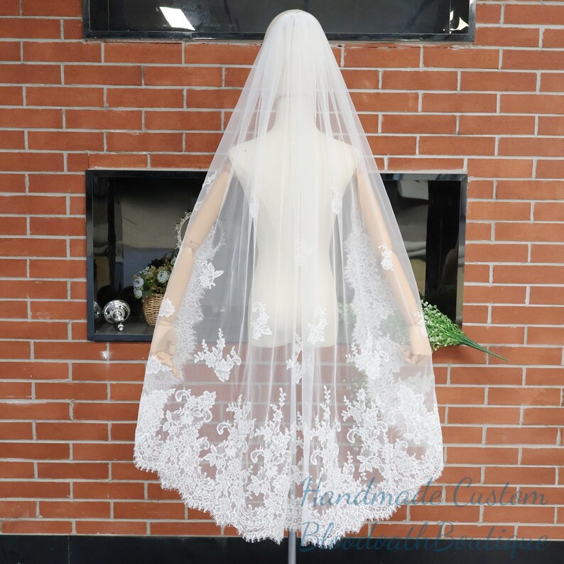 May include: A white bridal veil with intricate lace detailing along the edges. The veil is draped over a mannequin, showcasing its delicate design and flowing fabric. The background features a brick wall and a window.