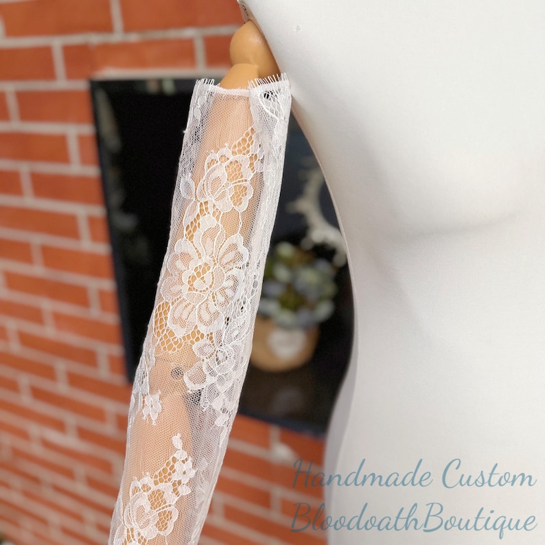May include: White lace sleeve with floral patterns, displayed on a mannequin arm. The delicate lace is semi-sheer, revealing the skin beneath. The sleeve is likely part of a bridal or formal wear ensemble.