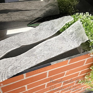 May include: White lace fabric strips with floral patterns are displayed on a dark surface. The lace is arranged in a layered fashion, with a hint of black lace visible. The background includes greenery and a brick wall.