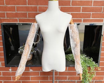May include: A pair of white lace sleeves on a mannequin. The sleeves are see-through, featuring a floral design. The mannequin is on a metal stand, set against a brick wall and a mirrored backdrop. These are bridal accessories.