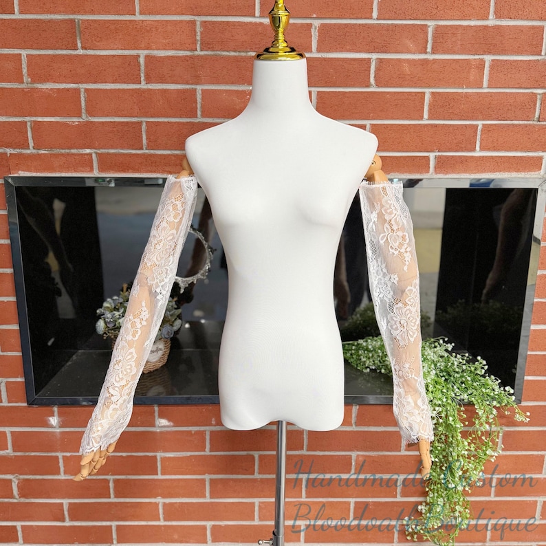 May include: White lace sleeves displayed on a mannequin. The sleeves are sheer, with a floral pattern. The mannequin is on a metal stand, set against a brick wall and a mirrored background. The sleeves are a bridal accessory.