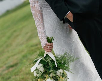 May include: A white lace sleeve and a white lace dress paired with a bouquet of white flowers and green foliage tied with a cream ribbon, alongside a black suit jacket. The bouquet features white blossoms and various green leaves.