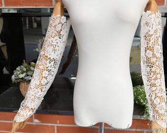 May include: A pair of white lace sleeves, featuring detailed floral patterns, are displayed on a mannequin. The sleeves are made of delicate, openwork lace. The mannequin is white, and the background includes brickwork and a floral arrangement.