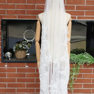 May include: A white bridal veil with intricate lace detailing cascading down the back. The veil is displayed on a mannequin against a red brick wall. The text "Handmade Custom Bloomworth Boutique" is visible.