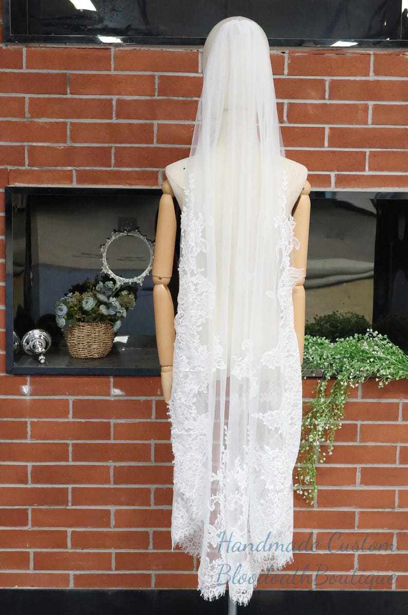 May include: A white bridal veil with intricate lace detailing cascading down the back. The veil is displayed on a mannequin against a red brick wall. The text "Handmade Custom Bloomworth Boutique" is visible.