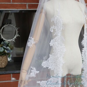 May include: A sheer white bridal veil with intricate white lace detailing along the edges. The veil is draped over a mannequin, showcasing the delicate floral patterns and the veil's flowing design. The text "Handmade Custom Bloodwash Boutique" is visible.