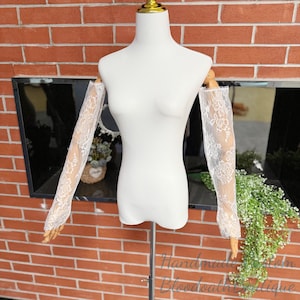 May include: White lace sleeves displayed on a mannequin. The sleeves are long, with a delicate floral pattern. The mannequin is white, and the background is a brick wall and a window. The text "Handmade from Bloodbath Boutique" is visible.