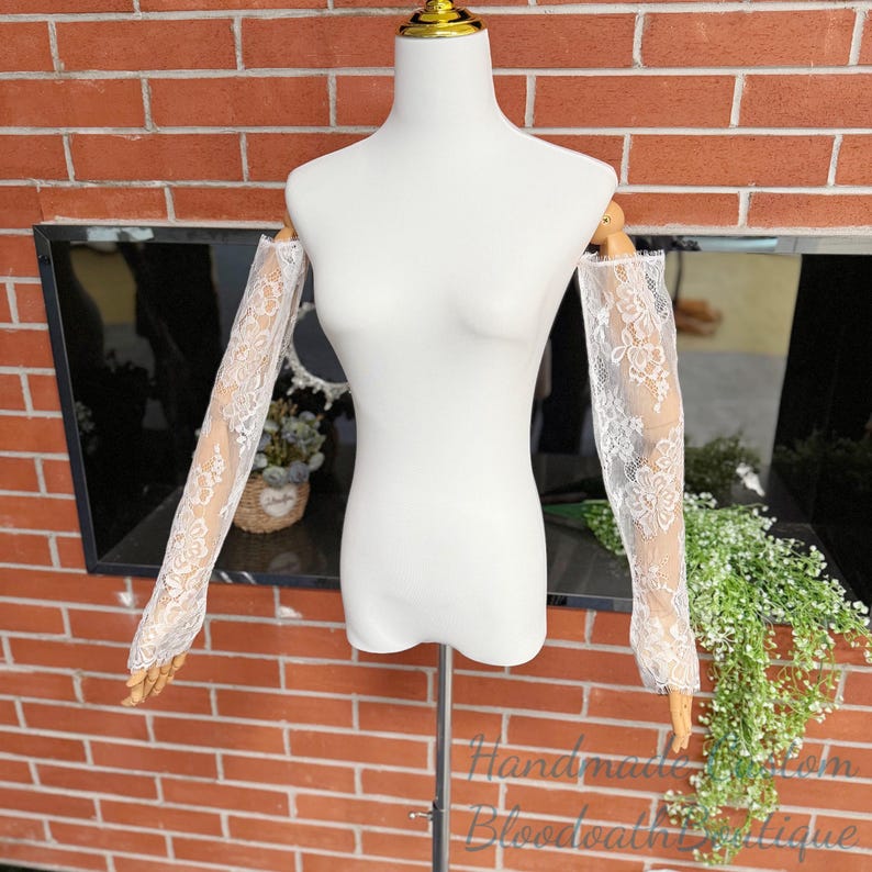 May include: White lace sleeves displayed on a mannequin. The sleeves are long, with a delicate floral pattern. The mannequin is white, and the background is a brick wall and a window. The text "Handmade from Bloodbath Boutique" is visible.