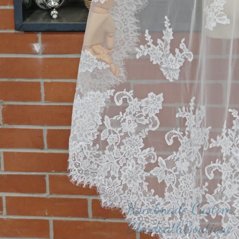 May include: A white lace veil with floral patterns. The delicate lace is draped, showcasing the intricate design and texture. The veil is against a brick wall background.