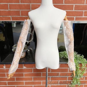 May include: White lace sleeves displayed on a mannequin. The sleeves are sheer, with intricate floral patterns. The mannequin is set against a brick wall background, with greenery and a mirror visible.