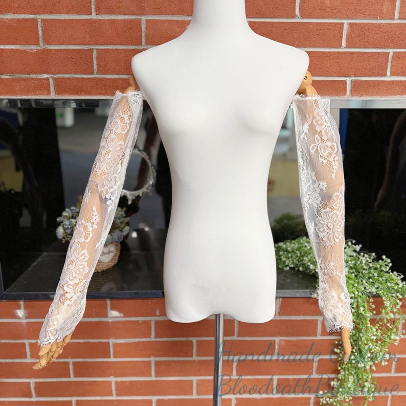May include: White lace sleeves displayed on a mannequin. The sleeves are sheer, with intricate floral patterns. The mannequin is set against a brick wall background, with greenery and a mirror visible.