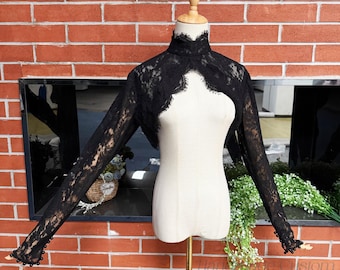 May include: Black lace bolero with long sleeves and a high, ruffled collar. The bolero is displayed on a mannequin against a brick wall background. The sleeves have a scalloped edge with small bead accents.