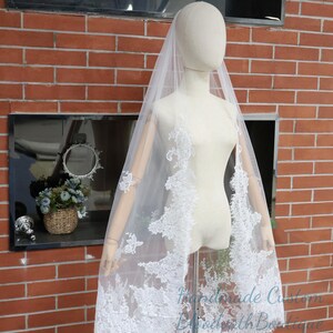 May include: A white bridal veil with intricate lace detailing. The veil is draped over a mannequin, showcasing the delicate floral and leaf patterns along the edges. The veil is sheer and lightweight, perfect for a wedding.