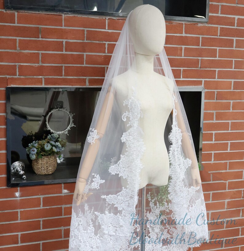 May include: A white bridal veil with intricate lace detailing. The veil is draped over a mannequin, showcasing the delicate floral and leaf patterns along the edges. The veil is sheer and lightweight, perfect for a wedding.