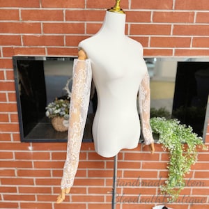 May include: A white dress form displays a pair of off-the-shoulder lace sleeves. The sleeves are made of delicate white lace with a floral pattern. The dress form is set against a red brick wall.