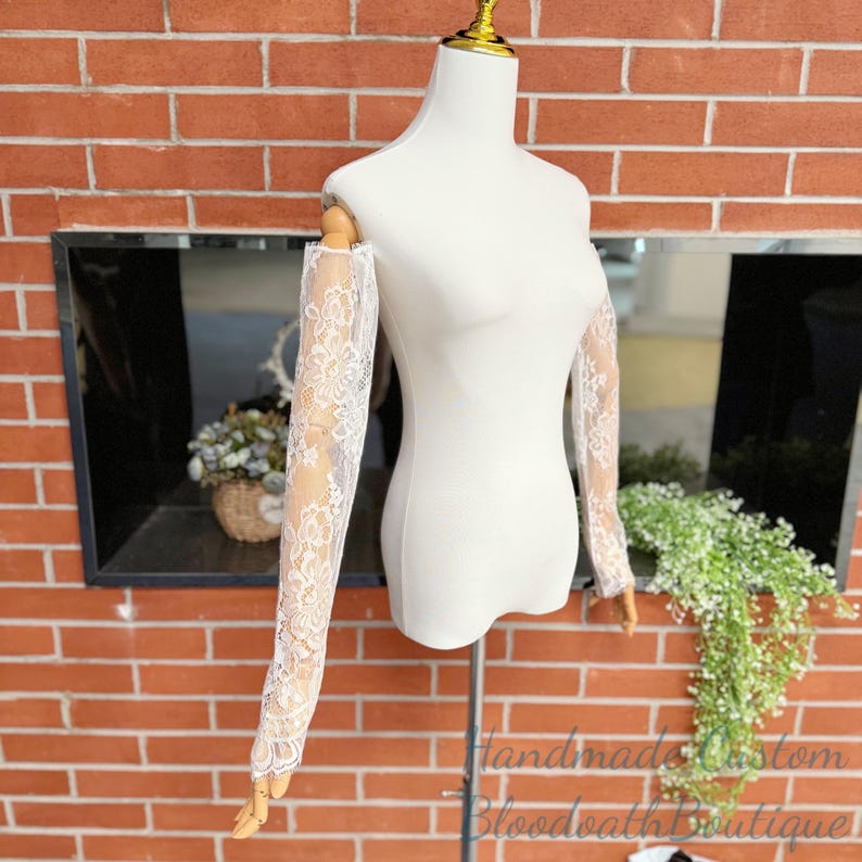 May include: A white dress form displays a pair of off-the-shoulder lace sleeves. The sleeves are made of delicate white lace with a floral pattern. The dress form is set against a red brick wall.