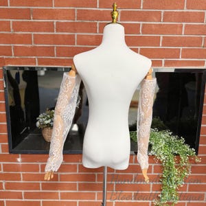 May include: White lace sleeves displayed on a mannequin. The sleeves are sheer, with a delicate floral pattern. The mannequin is white, and the background is a brick wall and a dark mirror.