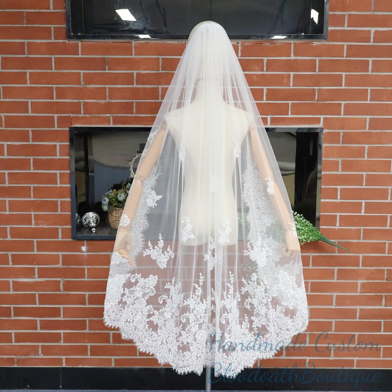 May include: A sheer white bridal veil with intricate floral lace detailing along the edges. The veil is draped over a mannequin, showcasing its delicate design and flowing fabric. The background features a brick wall.