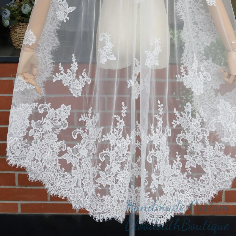 May include: A sheer white bridal cape with intricate floral lace detailing along the edges. The lace features delicate patterns of flowers and leaves, creating a romantic and elegant look. The cape is displayed on a mannequin, showcasing its flowing design.