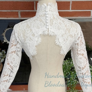 Può includere: Un bolero in pizzo bianco con maniche lunghe e colletto alto abbottonato. Il delicato design floreale in pizzo copre l'intero bolero, esposto su un manichino. La schiena presenta un design a goccia. Il testo "Handmade Bloodoa Boutique" è visibile.